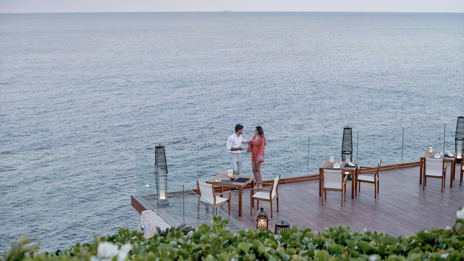 A man and woman talking on an outdoor terrace lined by glass railings, next to sea