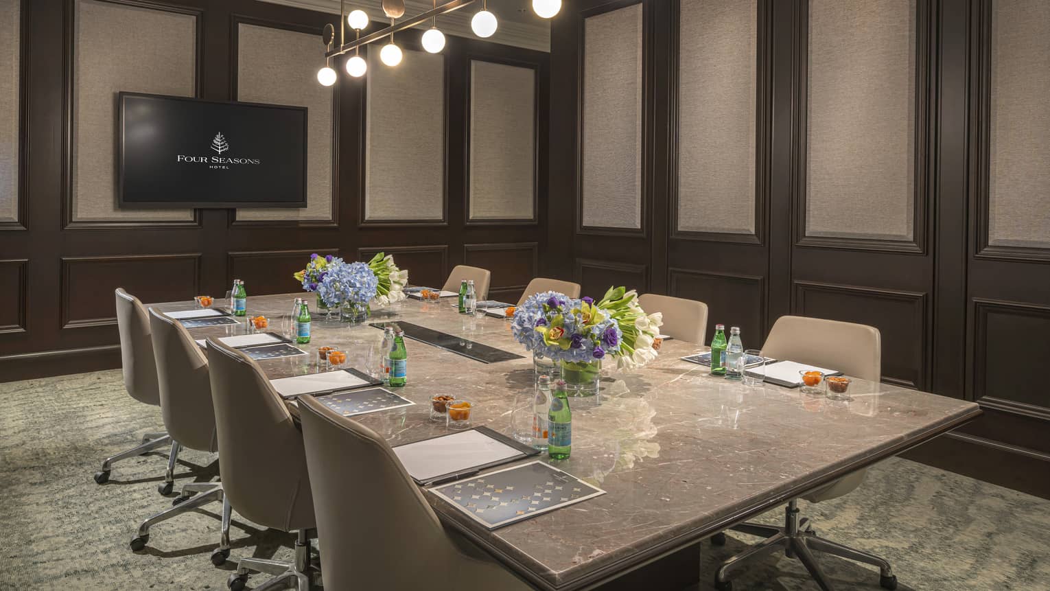 Meeting room with rectangular marble table and four chairs on either side, wall-mounted TV at front of room, green patterned carpet and modern chandelier