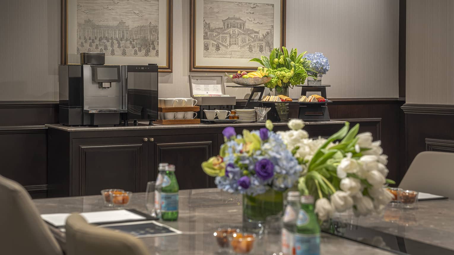 Marbled table with floral centrepiece in foreground, coffee station and fruit on sideboard in background of meeting room