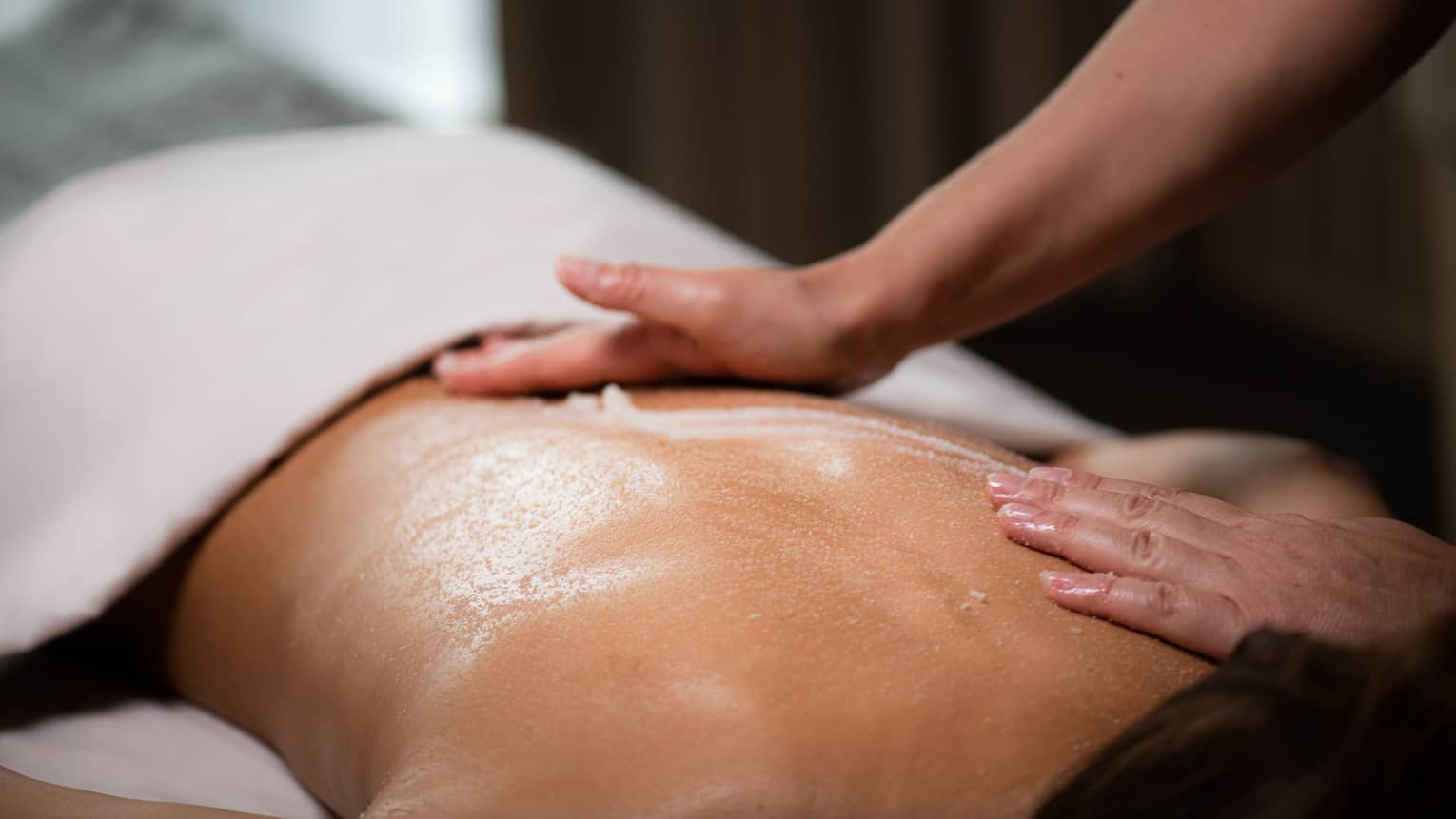 A masseuse spreads a cream across a woman's back as she lies on a massage table.