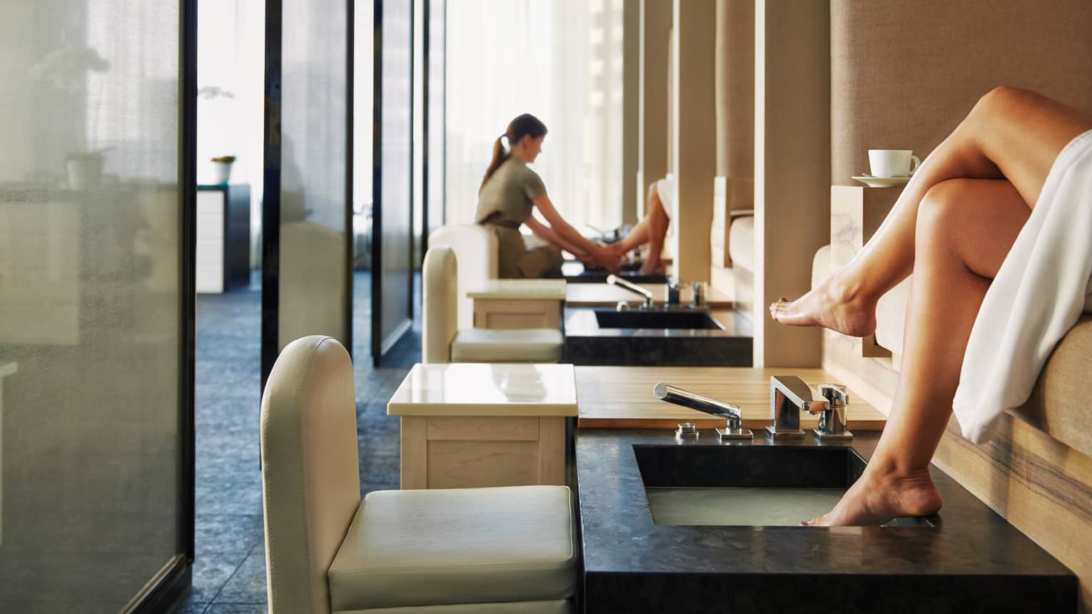 Woman's feet on pedicure sink along row of spa chairs, salon staff in background