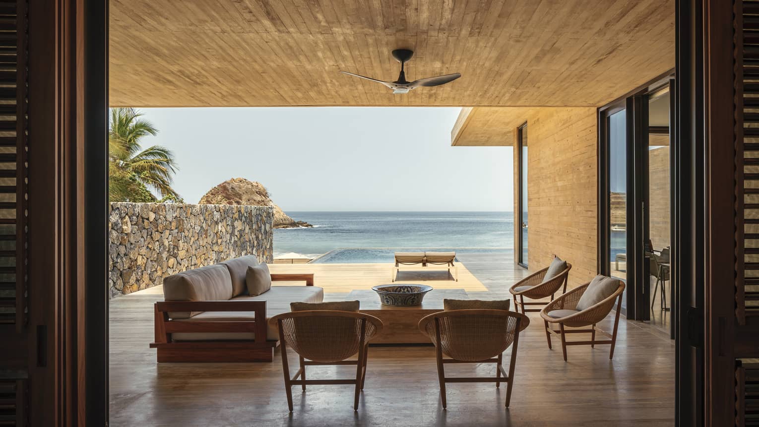 An open-air luxury villa overlooking the ocean, featuring a shaded seating area with modern wooden furniture and a private infinity pool extending towards the beach