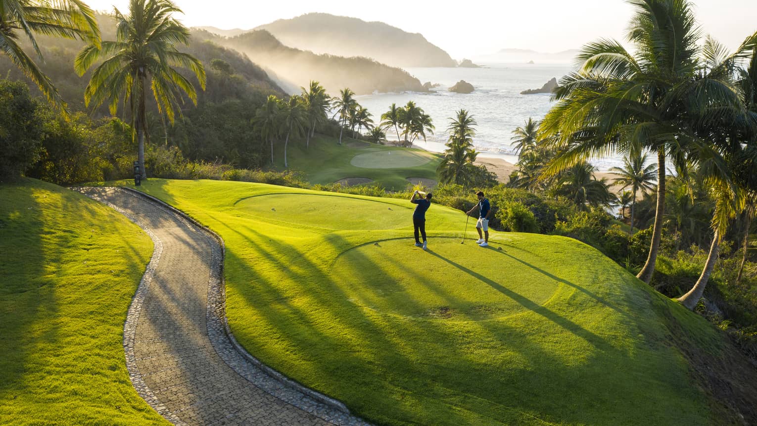 Rear view of two golfers on an elevated tee, one swinging toward the green down by the beach, misty hills and ocean beyond.