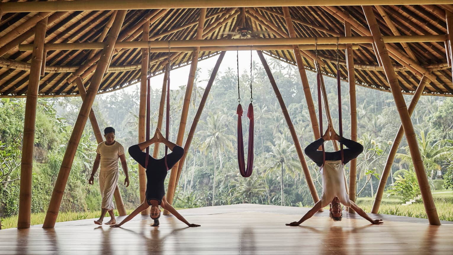 Woman and man hang upside-down on yoga hammocks under wood pavilion as instructor looks on
