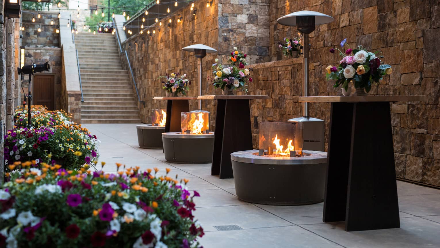 Outdoor event space with stone walls, string lights, floral arrangements on tall tables and firepits creating a warm ambience
