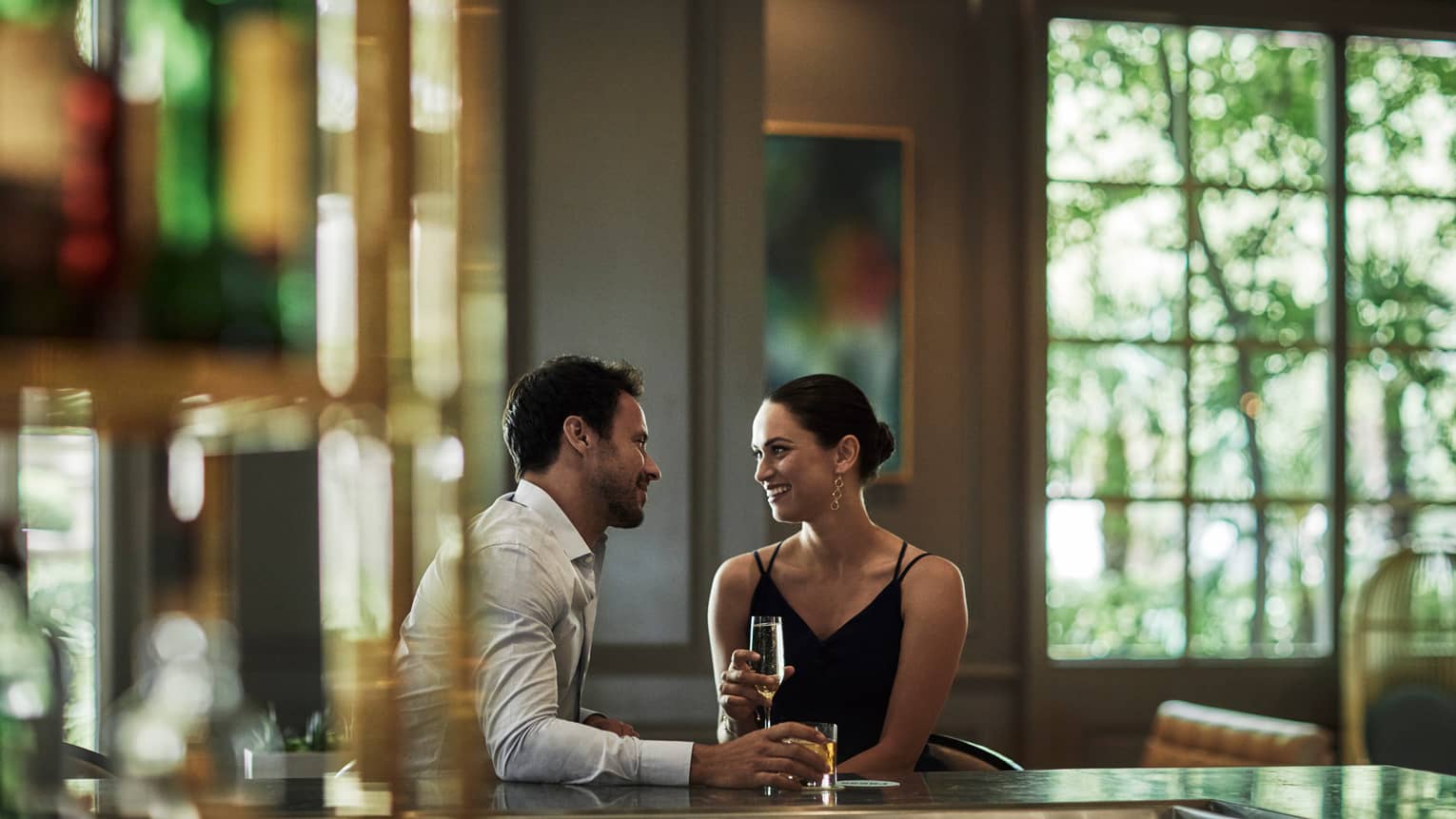 A man and woman talk at an indoor bar, window looking out to trees