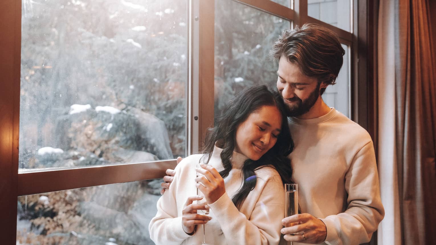 Man and woman embracing next to a window, holding champagne glasses