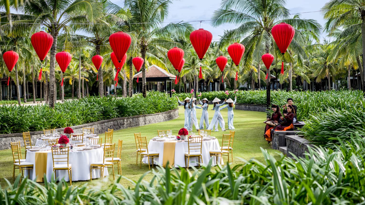 Women in traditional dress dance, accompanied by musicians, surrounded by red lanterns and palm trees