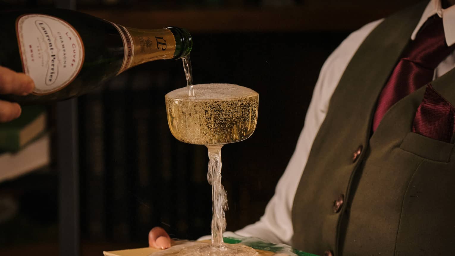 Person holding a stack of books with a champagne glass on top while another person pours champagne into the glass.