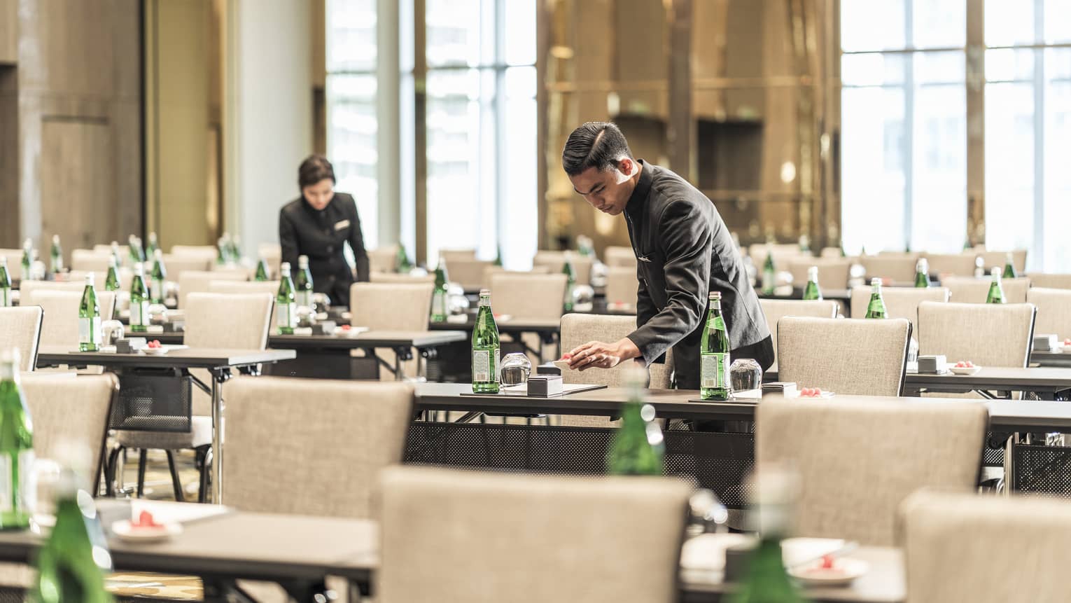 Hotel staff set agendas, water bottles on rows of meeting tables in conference ballroom