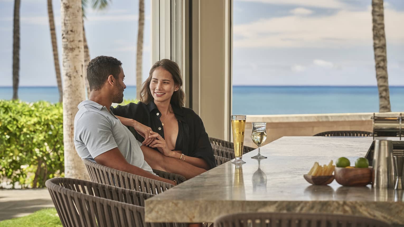 Couple sitting, smiling while seated at open-air bar with ocean in backdrop