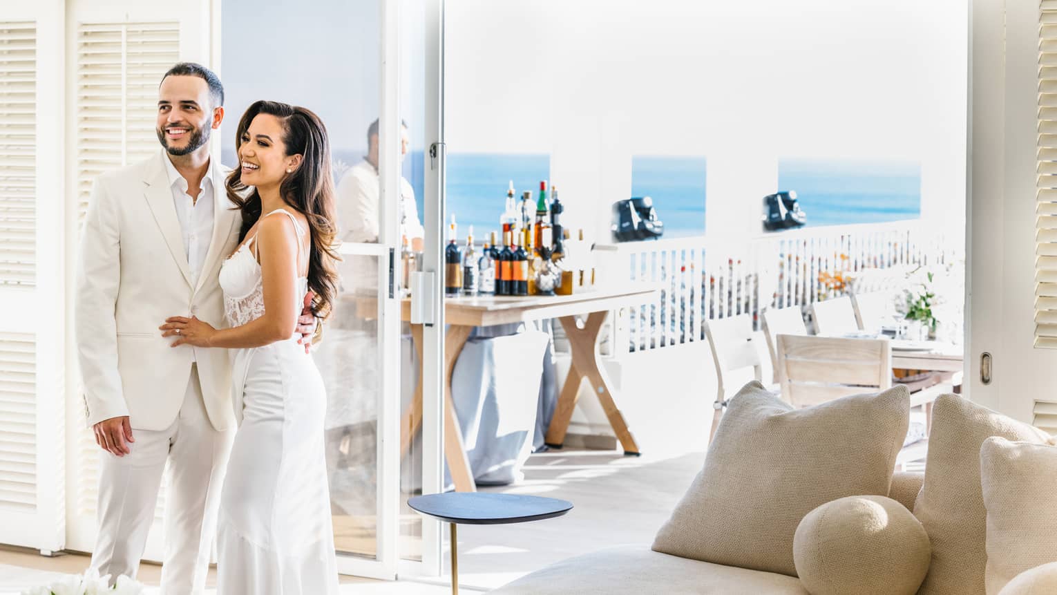 Groom and bride standing, smiling near terrace with bar setup and ocean view