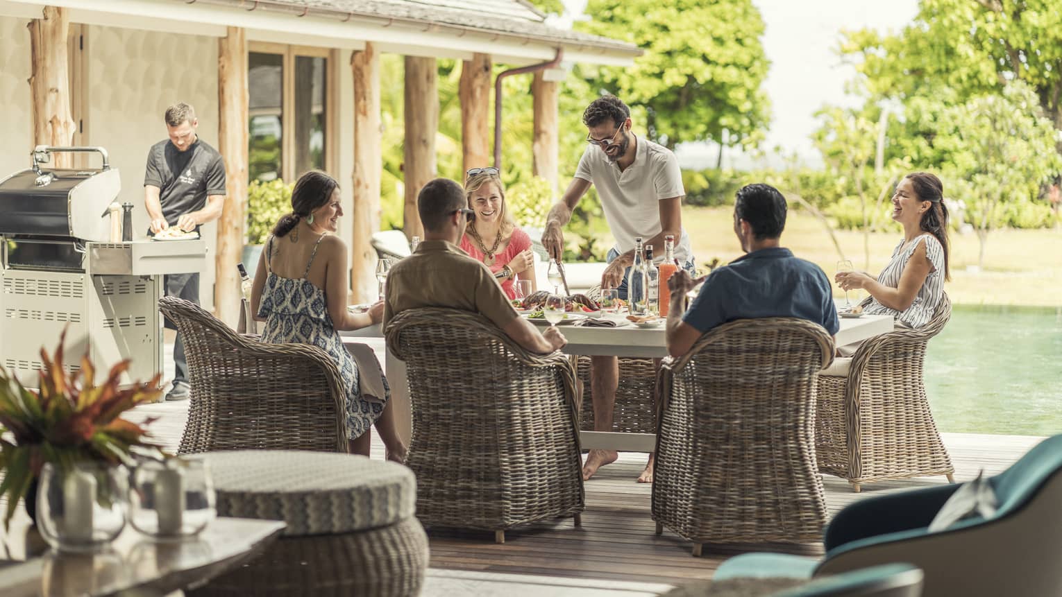 Group of friends enjoying an outdoor meal together on a sunny patio, with a chef preparing food on a grill nearby, creating a relaxed and social atmosphere.