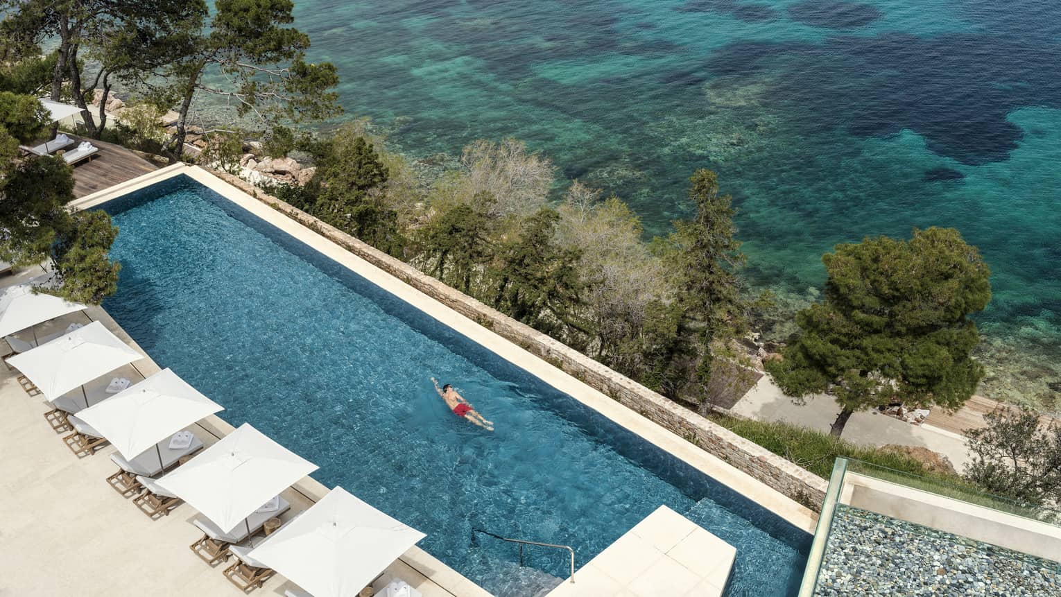 Man in red swim shorts swimming in infinity pool overlooking ocean and white cabanas 