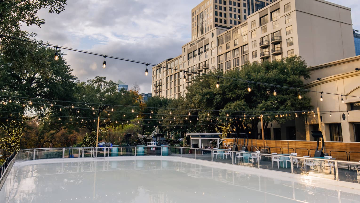 View of the Hotel overlooking an outdoor skating rink surrounded by trees and tables and lit by strings of Edison lights.
