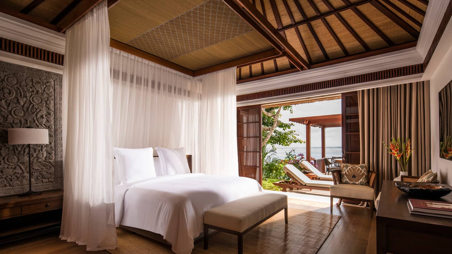 Villa bed with sheer white curtains hanging around each side, Balinese textiles on wall, door open to sunny patio