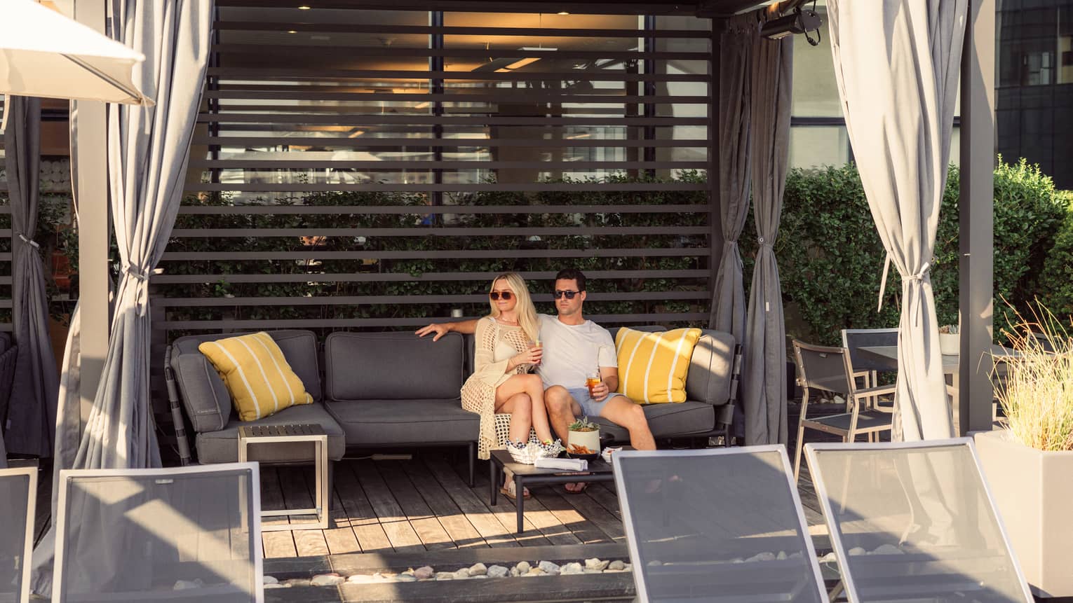 In a sun-dappled cabana, a couple relaxes together, drinks in hand, on a plush patio sofa, a table of snacks set for them.