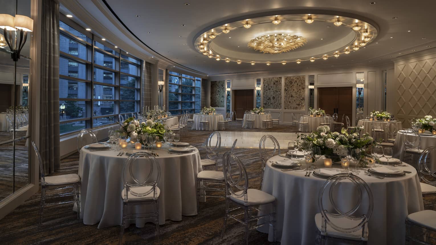 A dimly lit large meeting room with round tables covered in flowers and large windows.