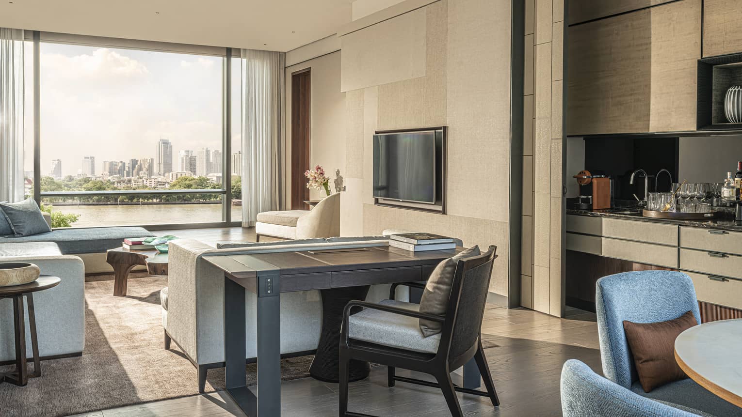 Hotel suite living area with blue, grey and beige furniture, console table, TV inset on beige wall, city view out large window