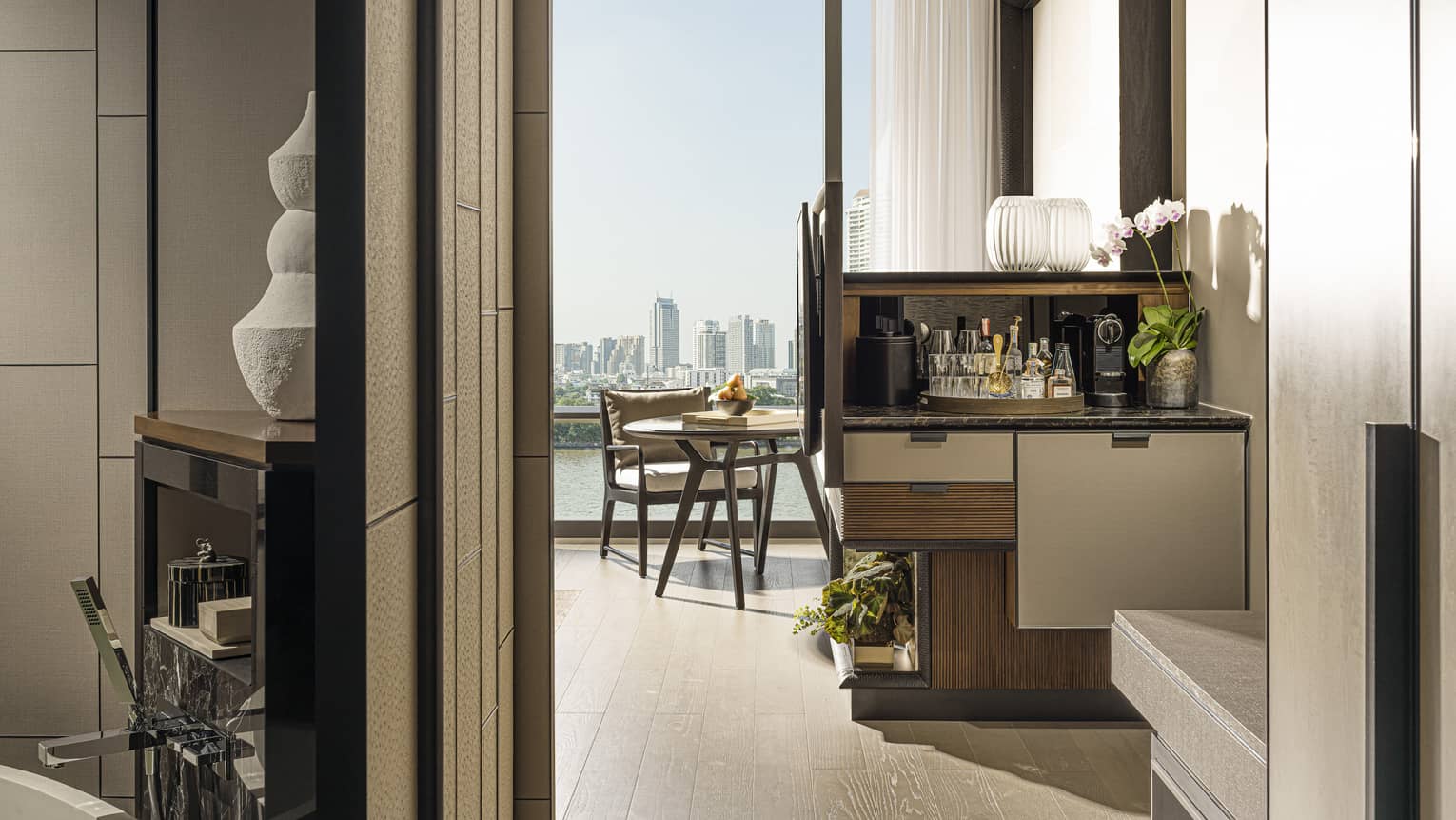 Hotel room entrance, glimpse of bathroom to left, wet bar, small dining table by tall window, city view