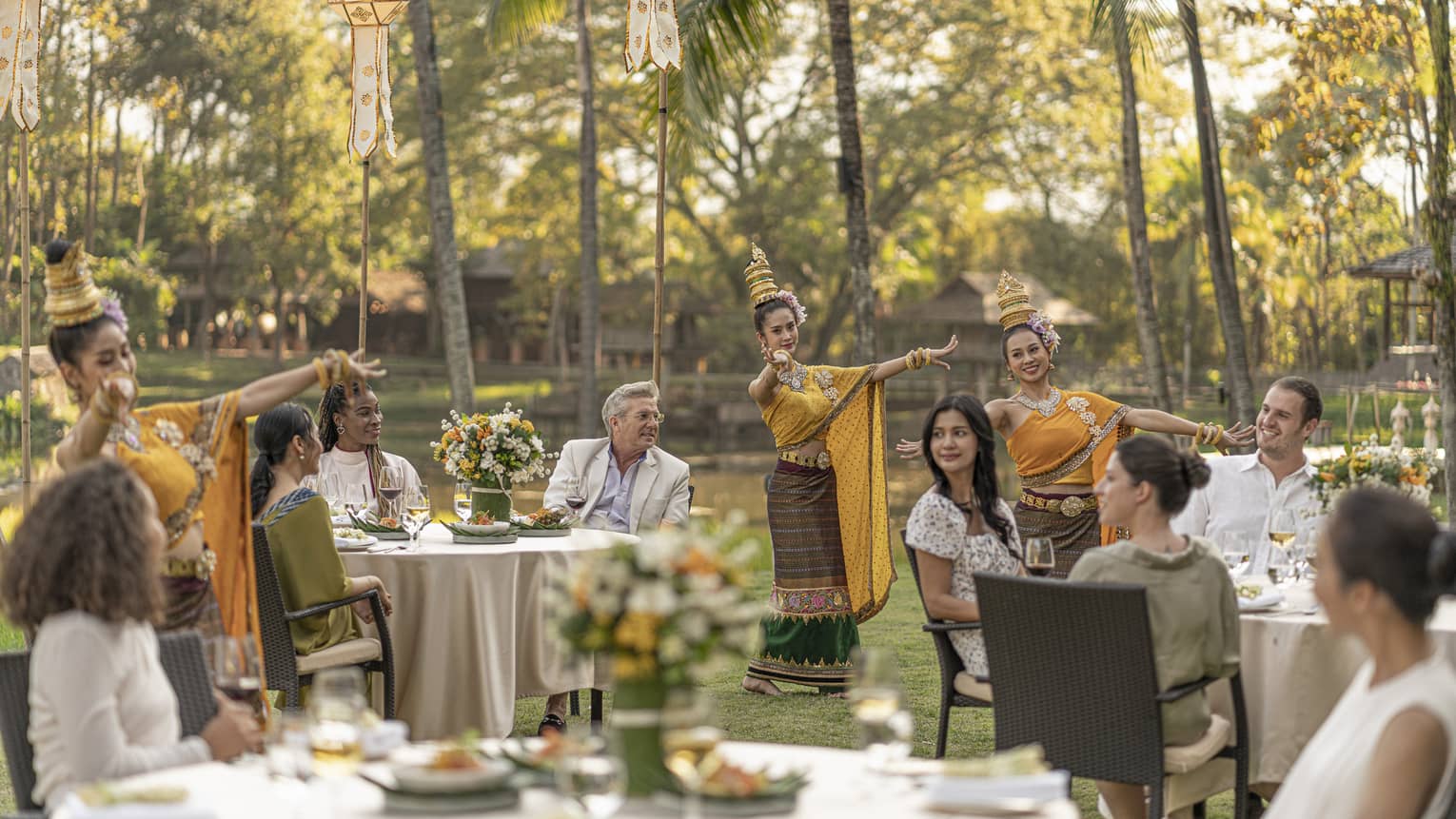 Several people sat in brown chairs, surrounded by dancers in orange robes beneath the palm trees.