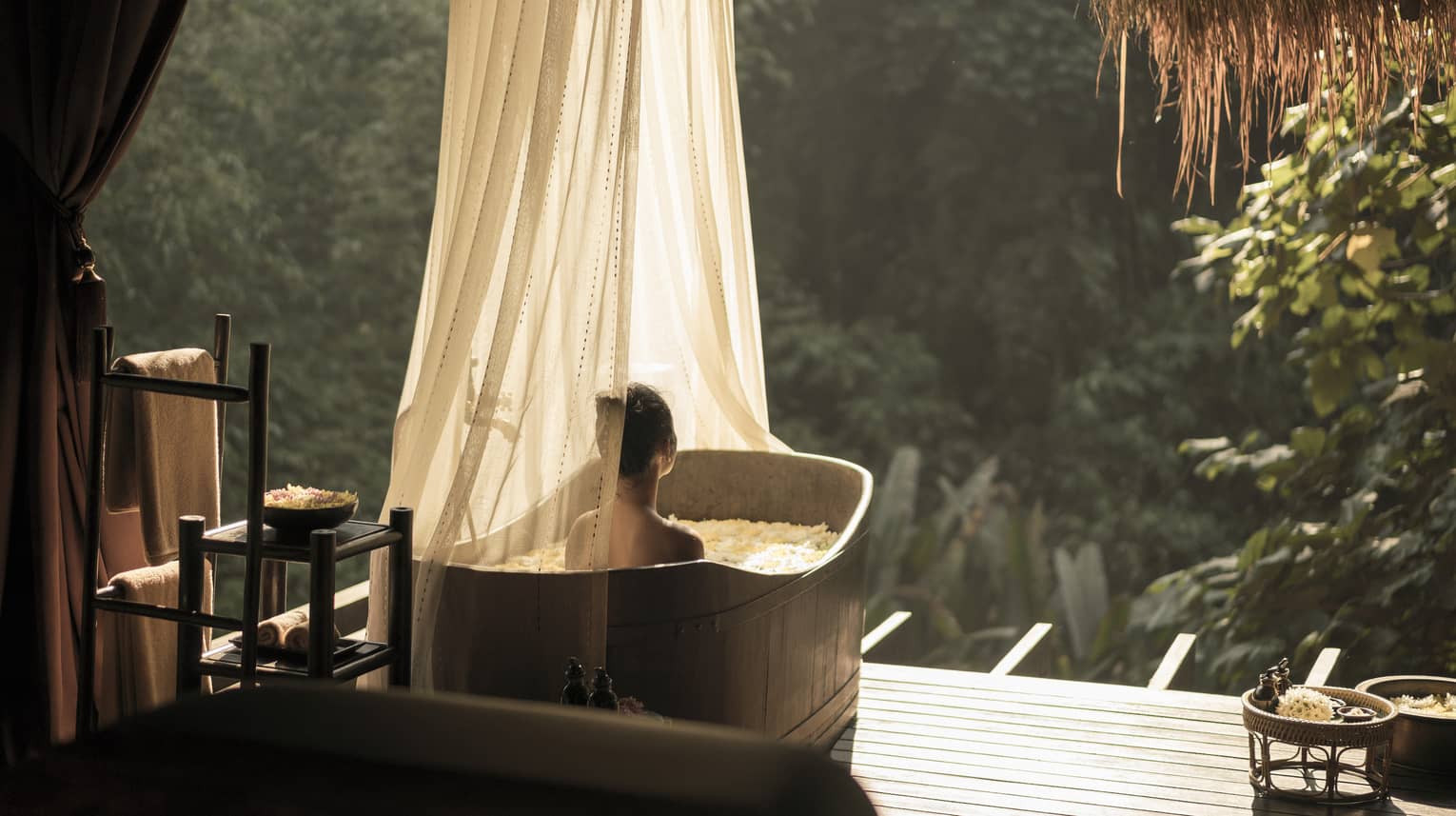 A person relaxes in an outdoor bathtub with sheer curtains, surrounded by lush greenery and sunlight. Towels and bath items are neatly arranged nearby, creating a serene spa atmosphere.