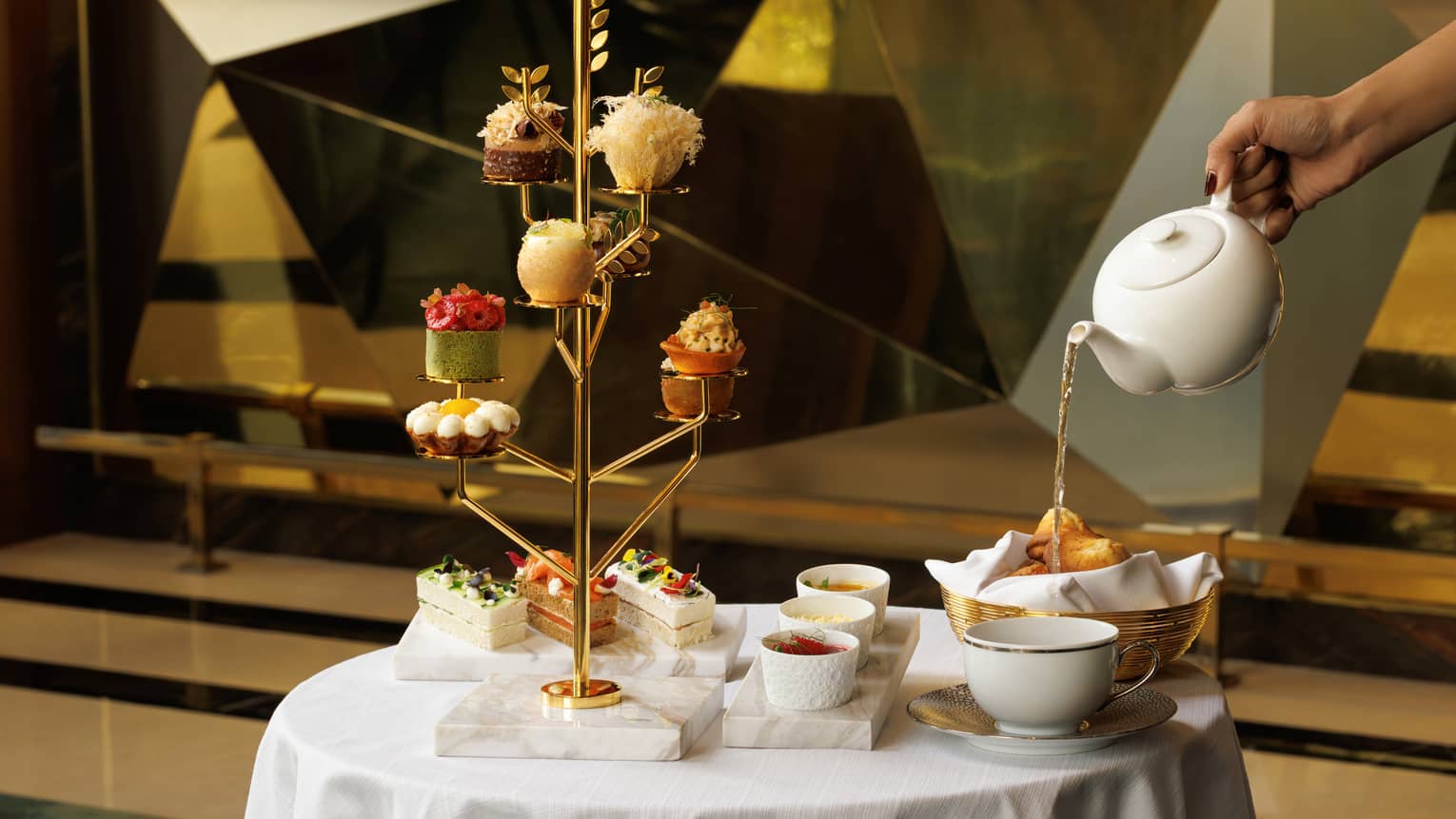 Table set for afternoon tea with a gold tree-shaped pastry stand, white marble platters with finger sandwiches and small bowls, a gold break basket and a hand reaching in pouring tea from a white teapot into a white mug