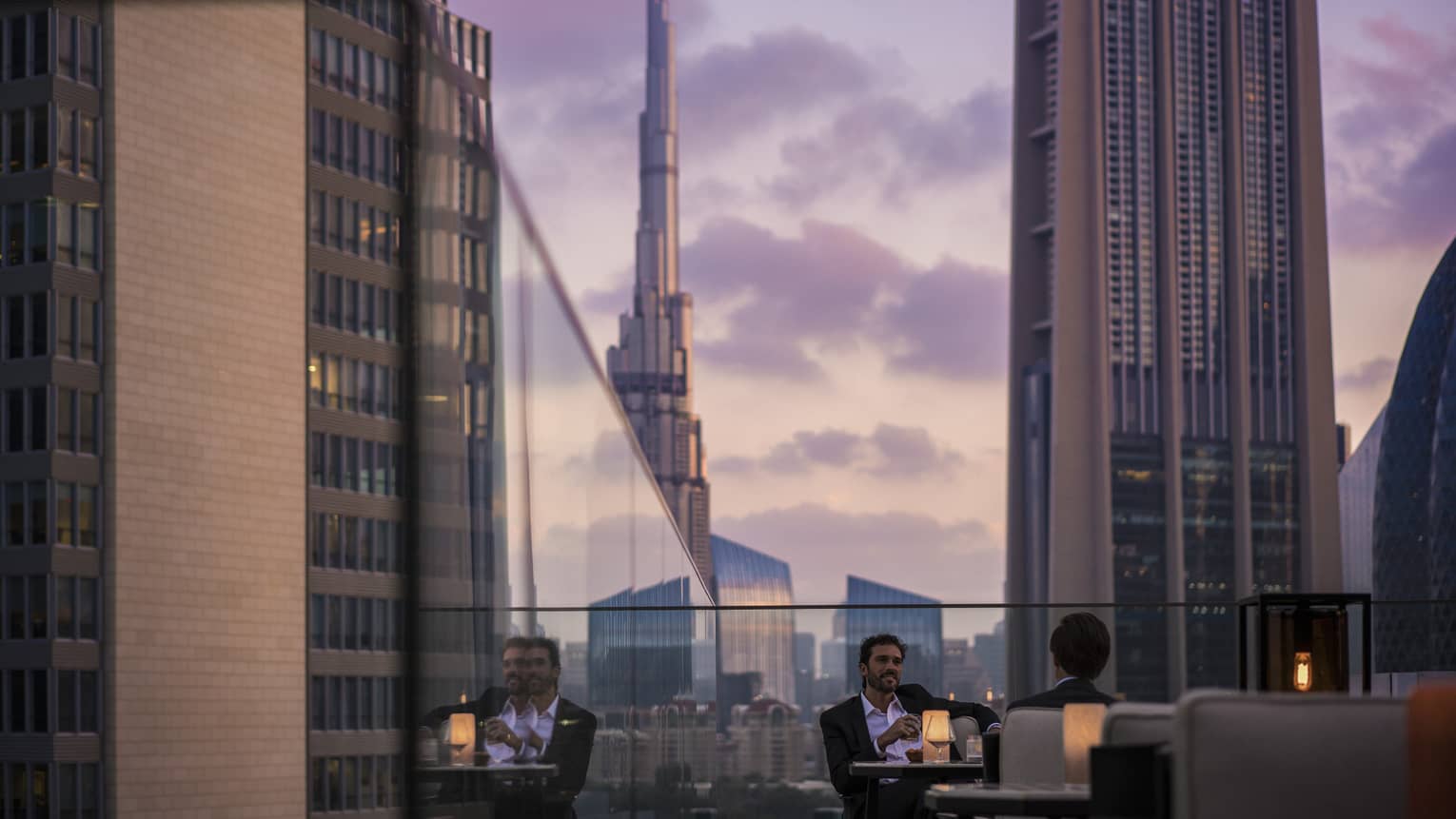 Men in suits on rooftop patio in front of glass balcony wall overlooking city at sunset