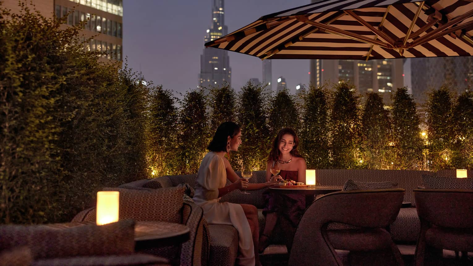 Two women sit at an outdoor table at dusk with a candle in the middle and a patio umbrella overhead