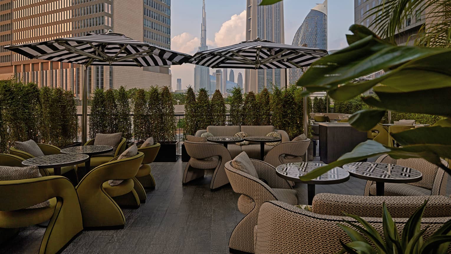 Outdoor restaurant patio filled with small bistro tables, beige upholstered chairs, black-and-white striped umbrellas and a hedge of bushes lining the ledge