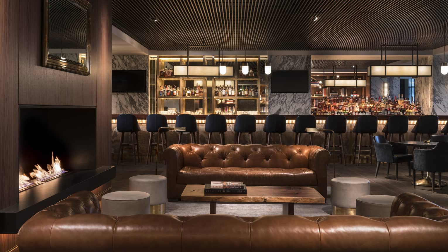 Two caramel-coloured Chesterfield sofas around a lit fireplace with cocktail bar in the background