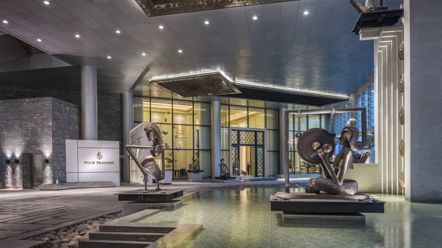 Hotel entrance with large modern Indonesian statues in fountain, doorman holding open glass doors in background