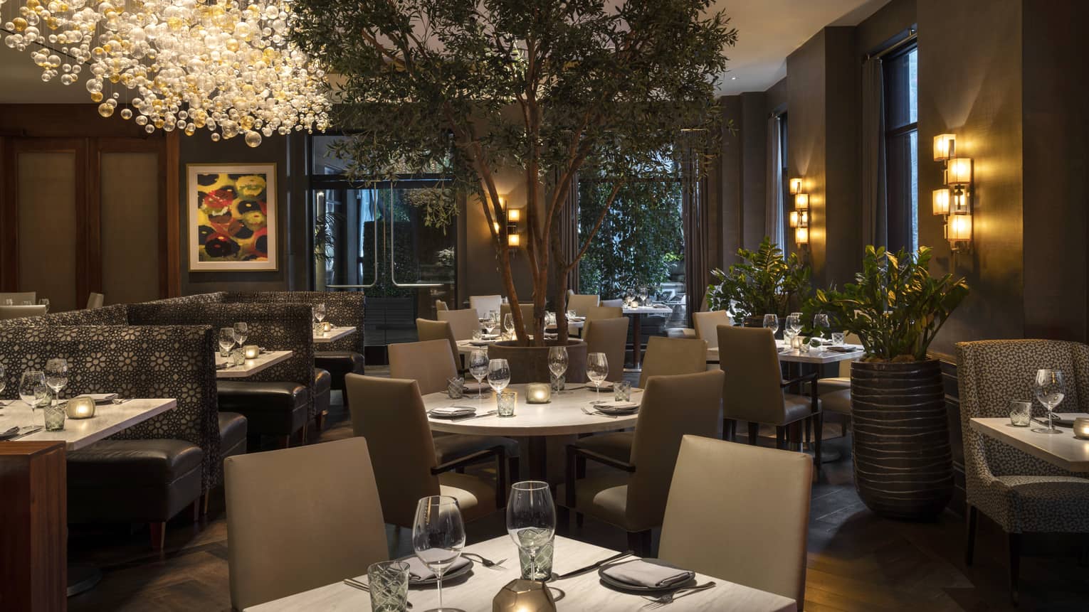 Indoor restaurant in the evening with square tables, taupe chairs, large tree, long hanging chandelier