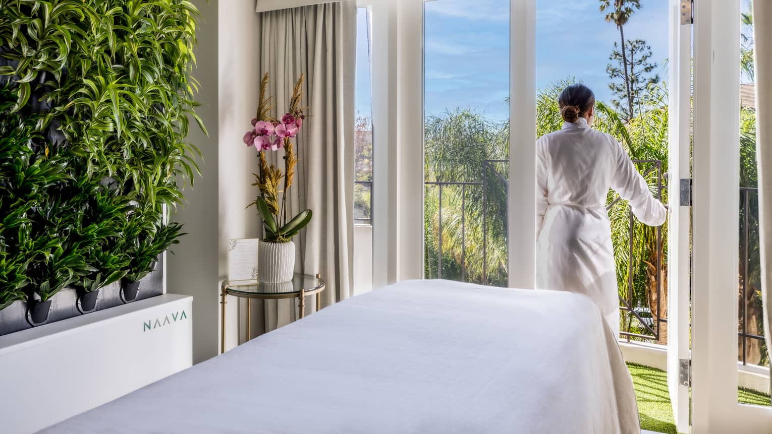 A woman walking outside from a massage room with large windowed doors and plants on one wall.