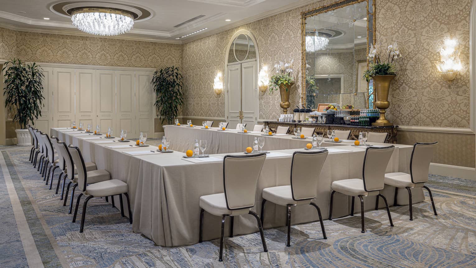 Meeting room set up hollow square with cream-coloured linens, dual chandeliers, lighted wall sconces and large mirror on patterned wall