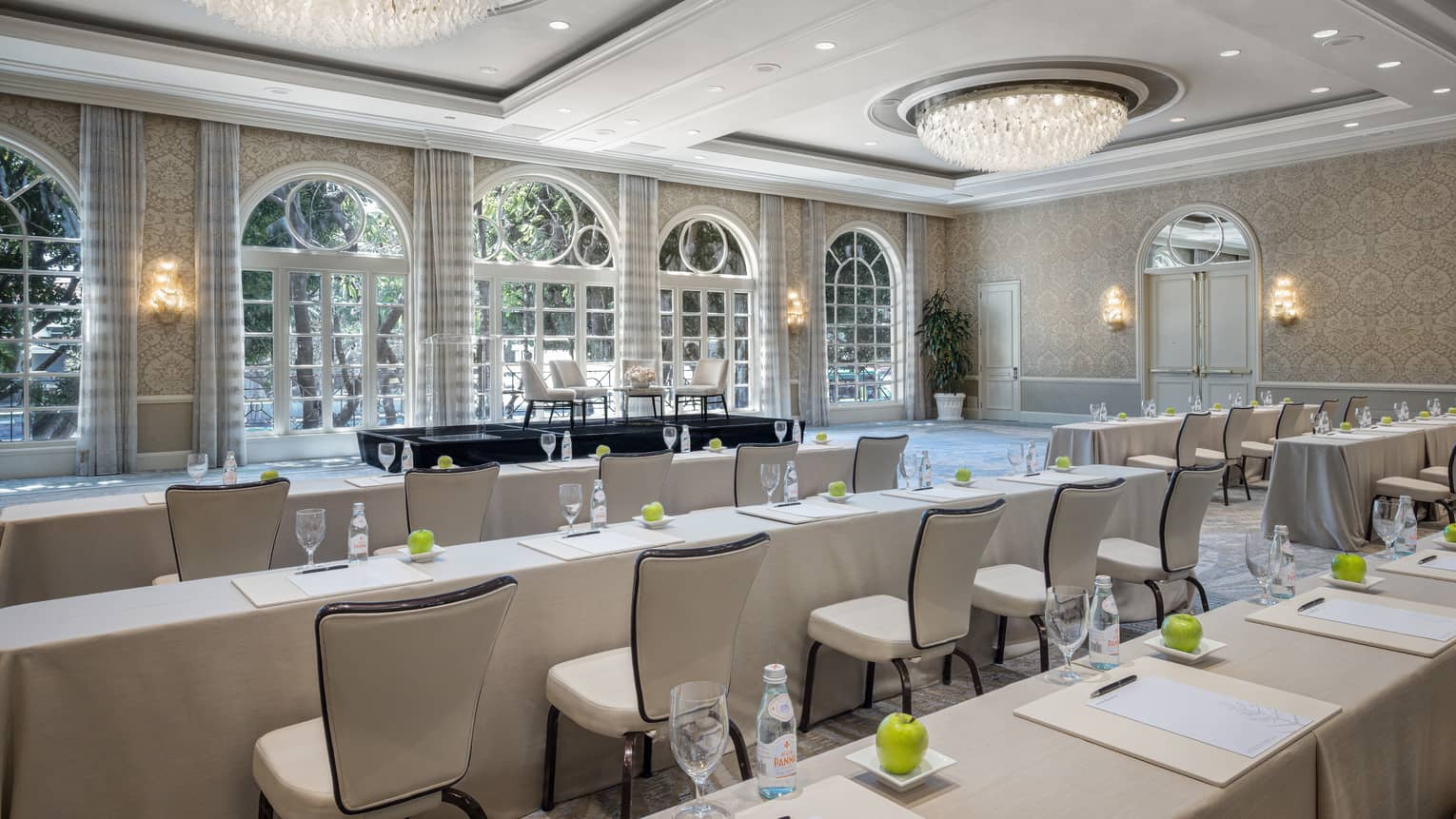 Ballroom set classroom style with cream-coloured linens, dual chandeliers, lighted wall sconces and floor-to-ceiling windows