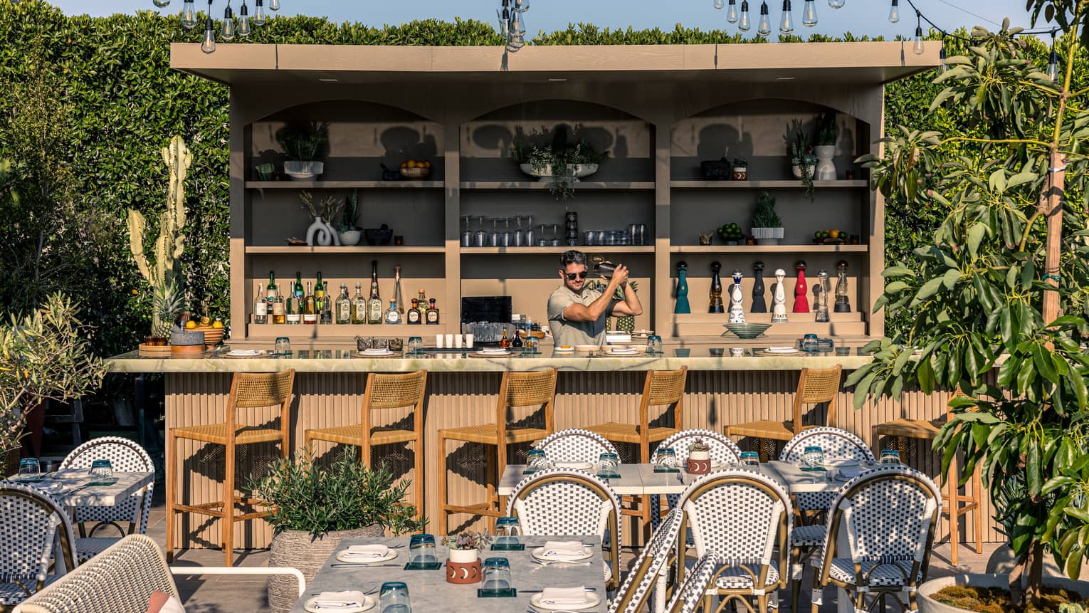 Outdoor restaurant patio with bartender standing behind a wood bar counter with shelving behind them and sting lights overhead