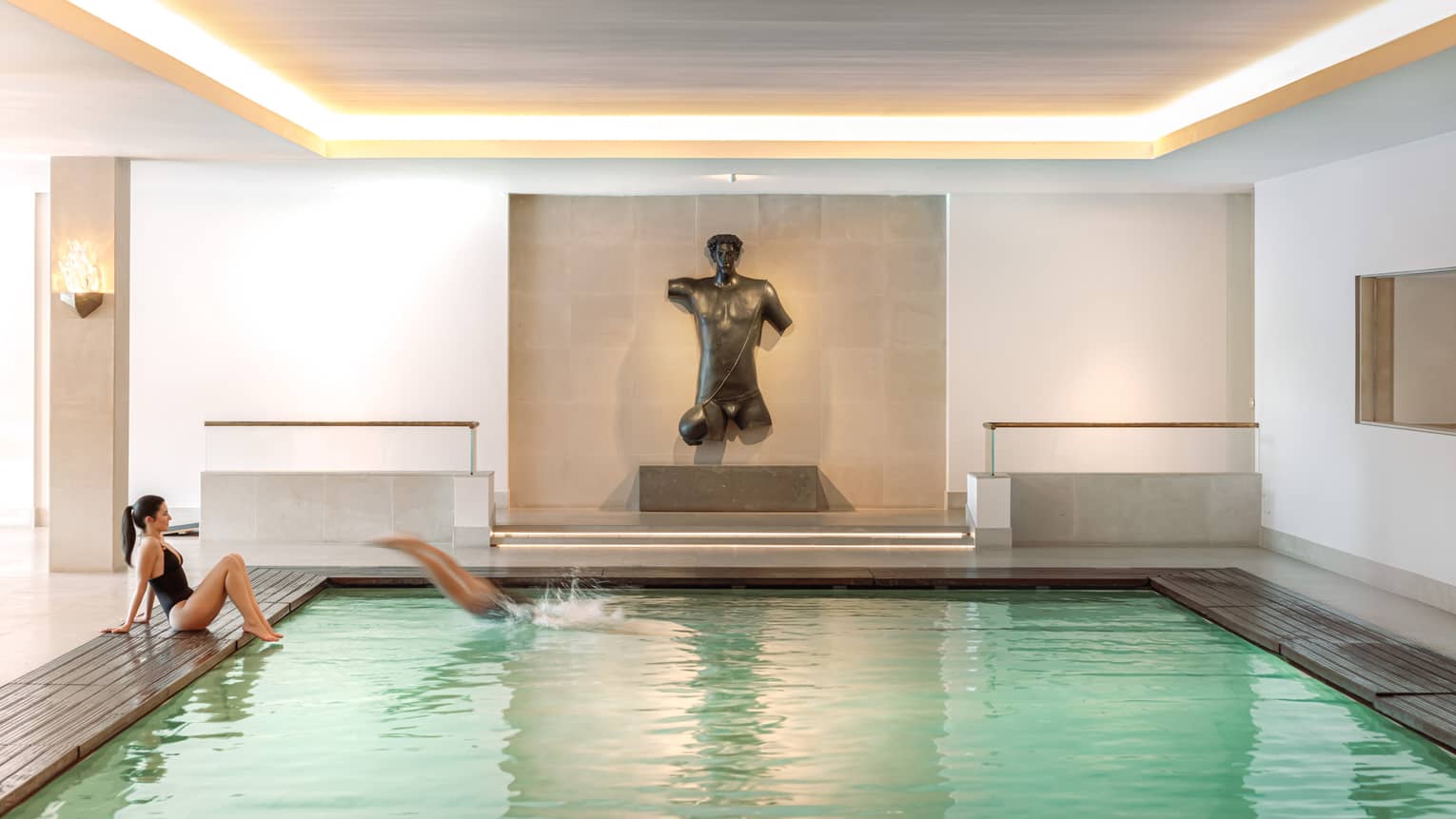 An indoor pool in a bright space finished in neutral tones. A figurative sculpture is displayed against a wall at the end of the pool.