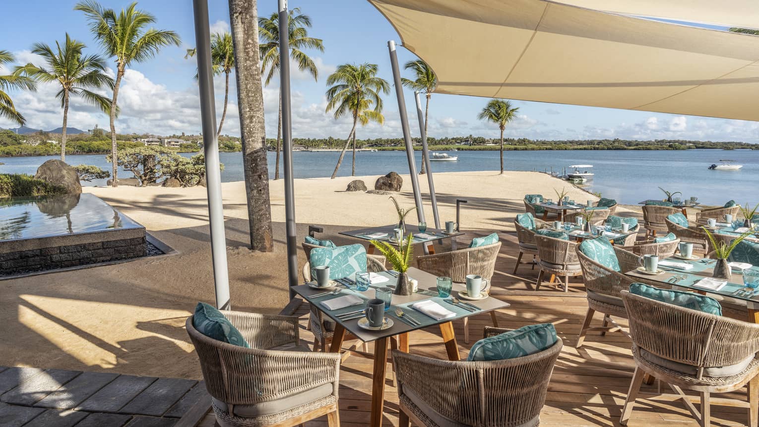 Wicker table and chair seating with aqua pillows under tan shade at waterfront