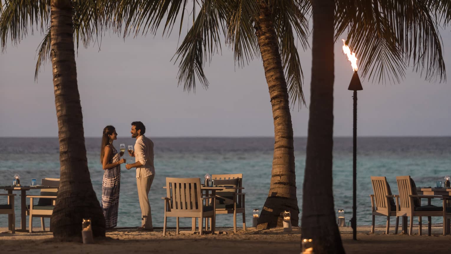 A couple talks on the beach at sunset, a tiki torch burns, table and chairs sit on beach