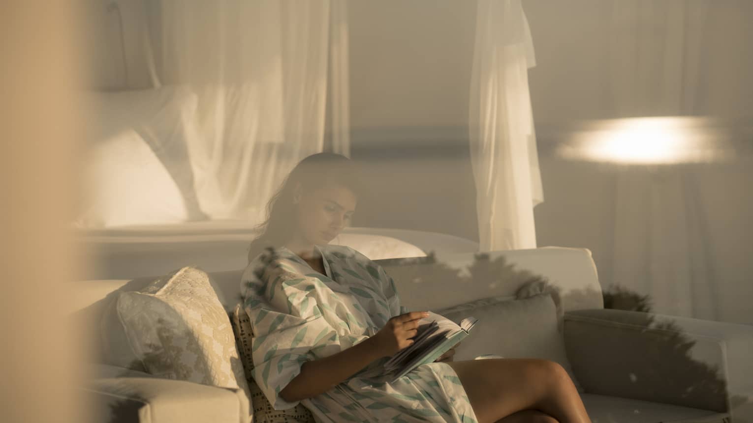 Woman in white robe with green print behind glass, reads book on white sofa