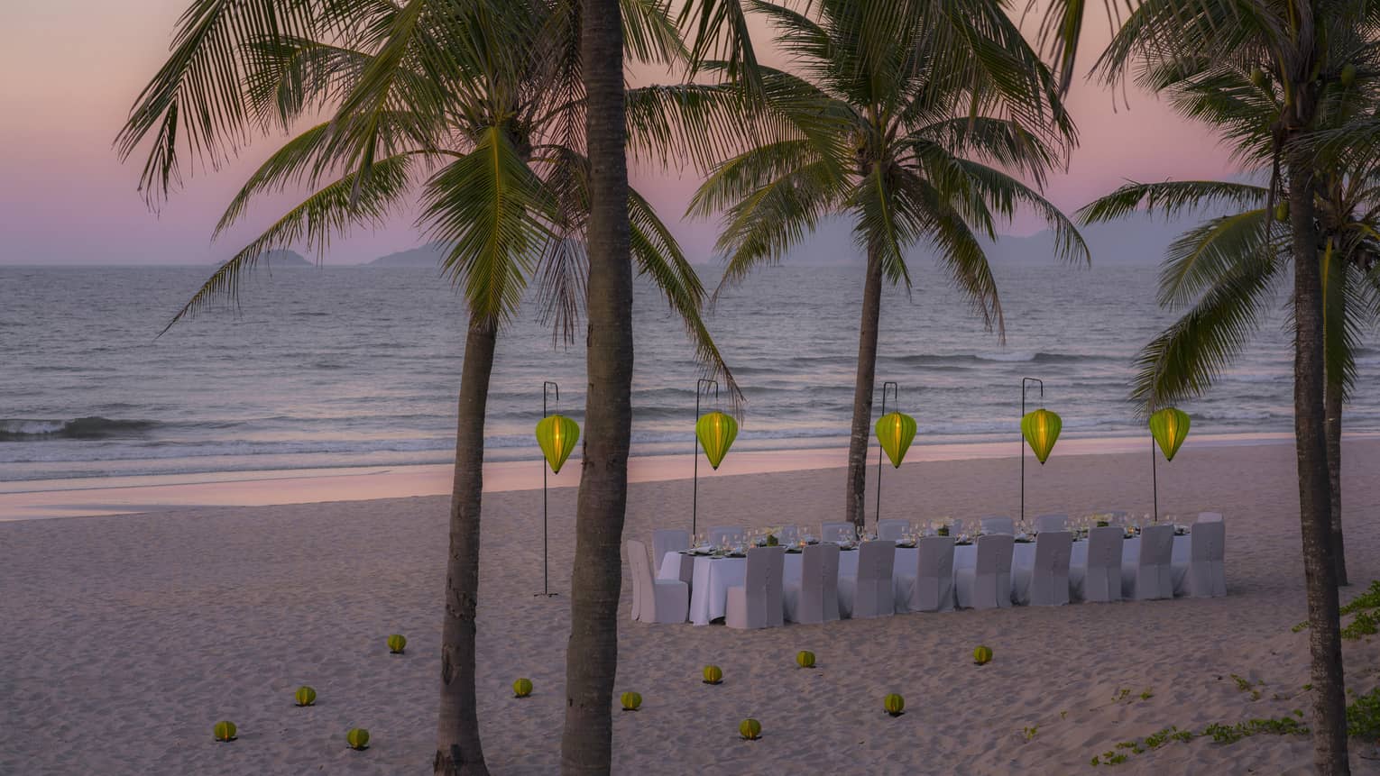 Palm trees and long dining table on beach with paper lanterns at sunset