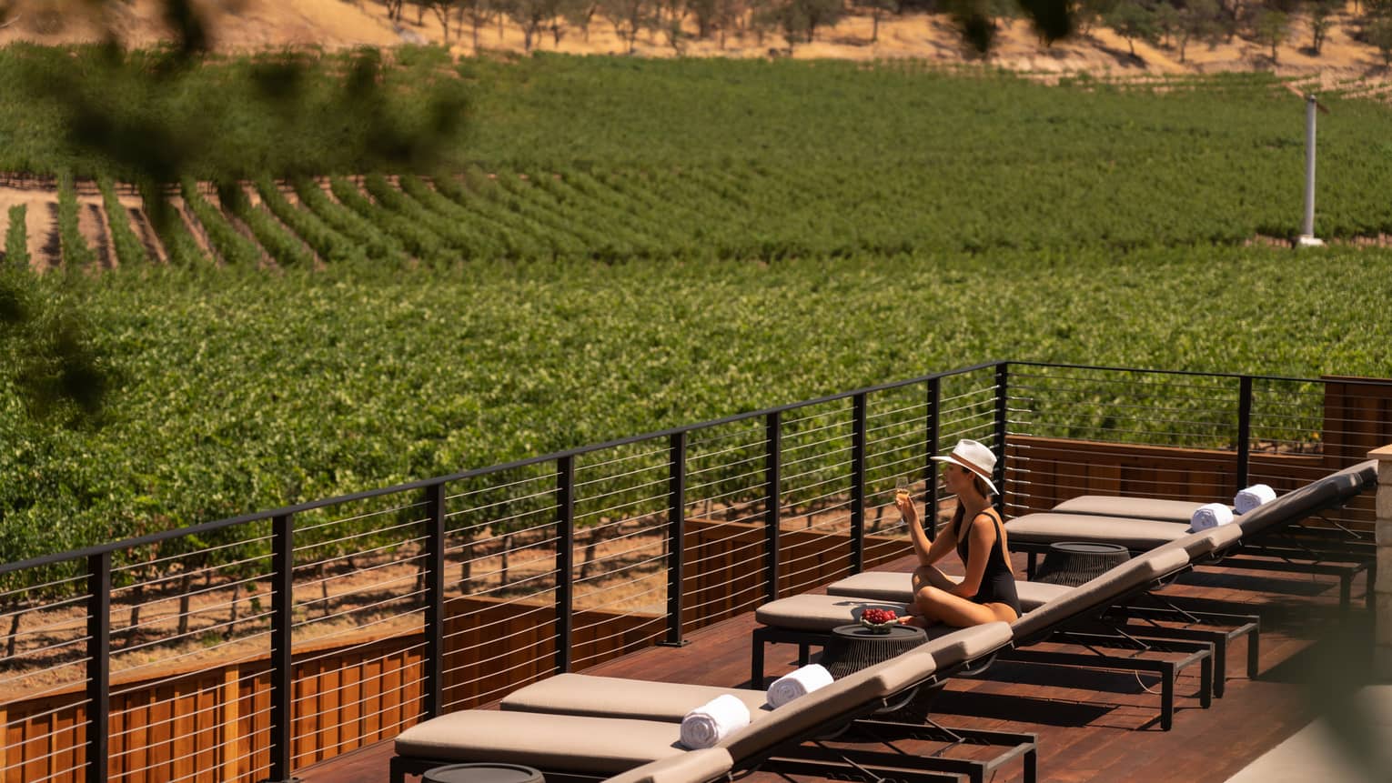 A person in a sunhat relaxing on a lounge chair with a glass of wine, overlooking a vast vineyard under the warm sunlight.