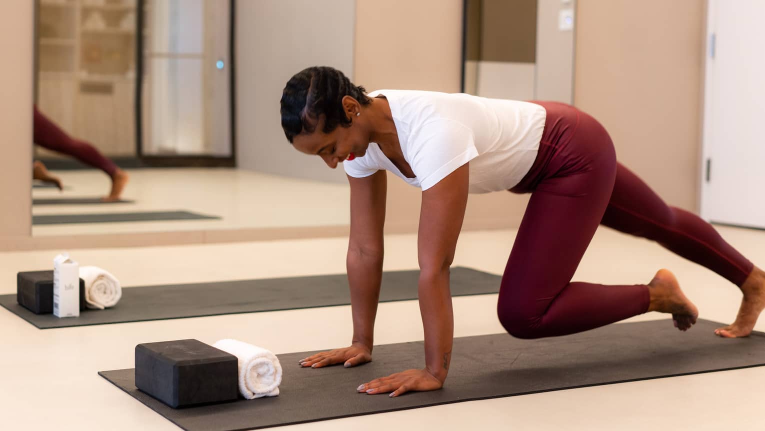 Person wearing burgundy leggings and a white short-sleeve t-shirt streches on a black yoga mat on a studio floor