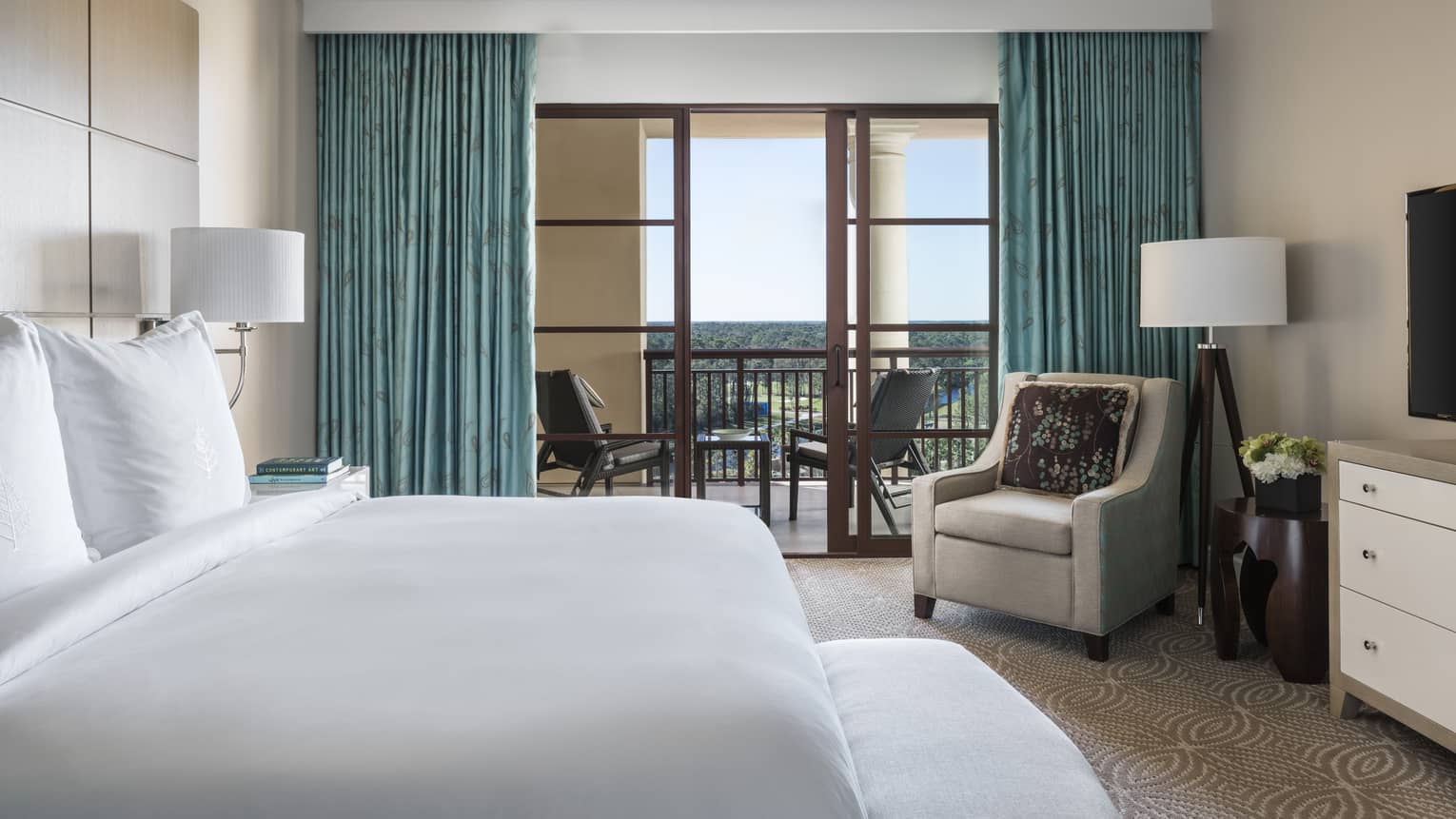 Park View Suite bed with white linens, bench, armchair by balcony windows with blue curtains