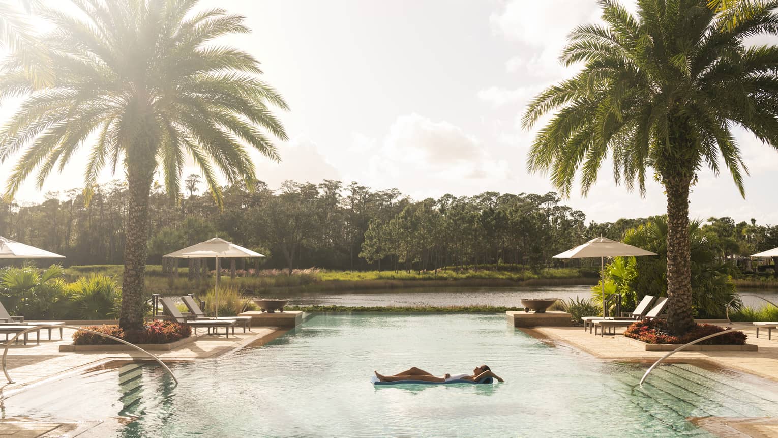 Swimmer floats in outdoor swimming pool under large palm trees