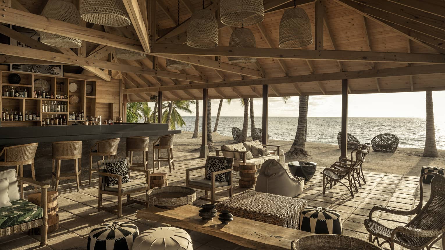 Bar with bar stools and lounge with grouped seating under vaulted wooden ceiling with open-air view of beach and ocean