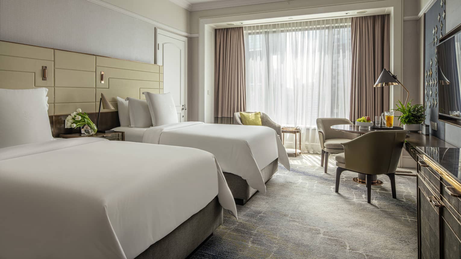 Guest room with two double beds, two-seater round table with arm chairs, and floor-to-ceiling window with beige curtains