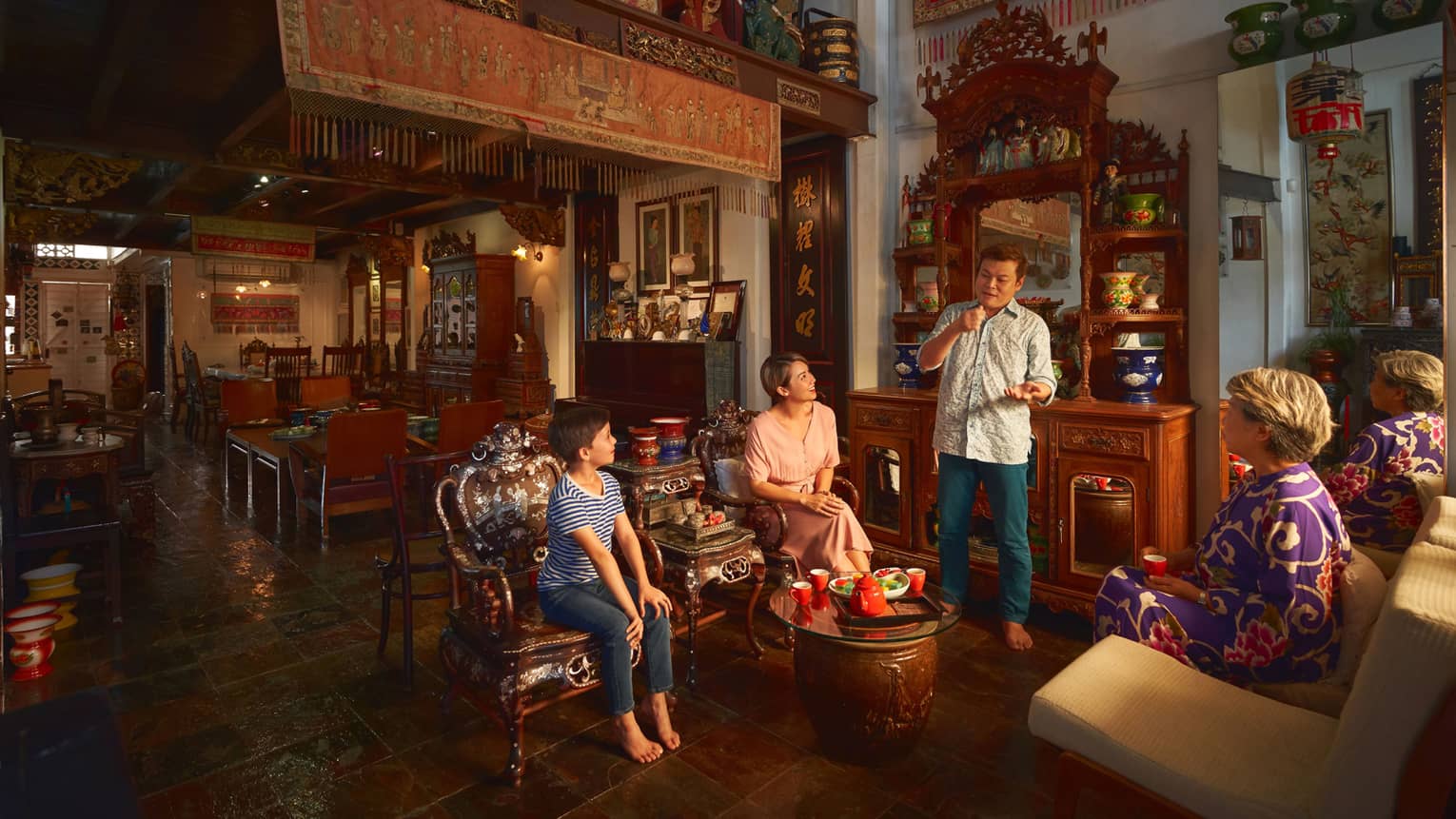 A family gathers around a low, round table holding a tea set in a room with ornate furniture, porcelain vases and tapestries.