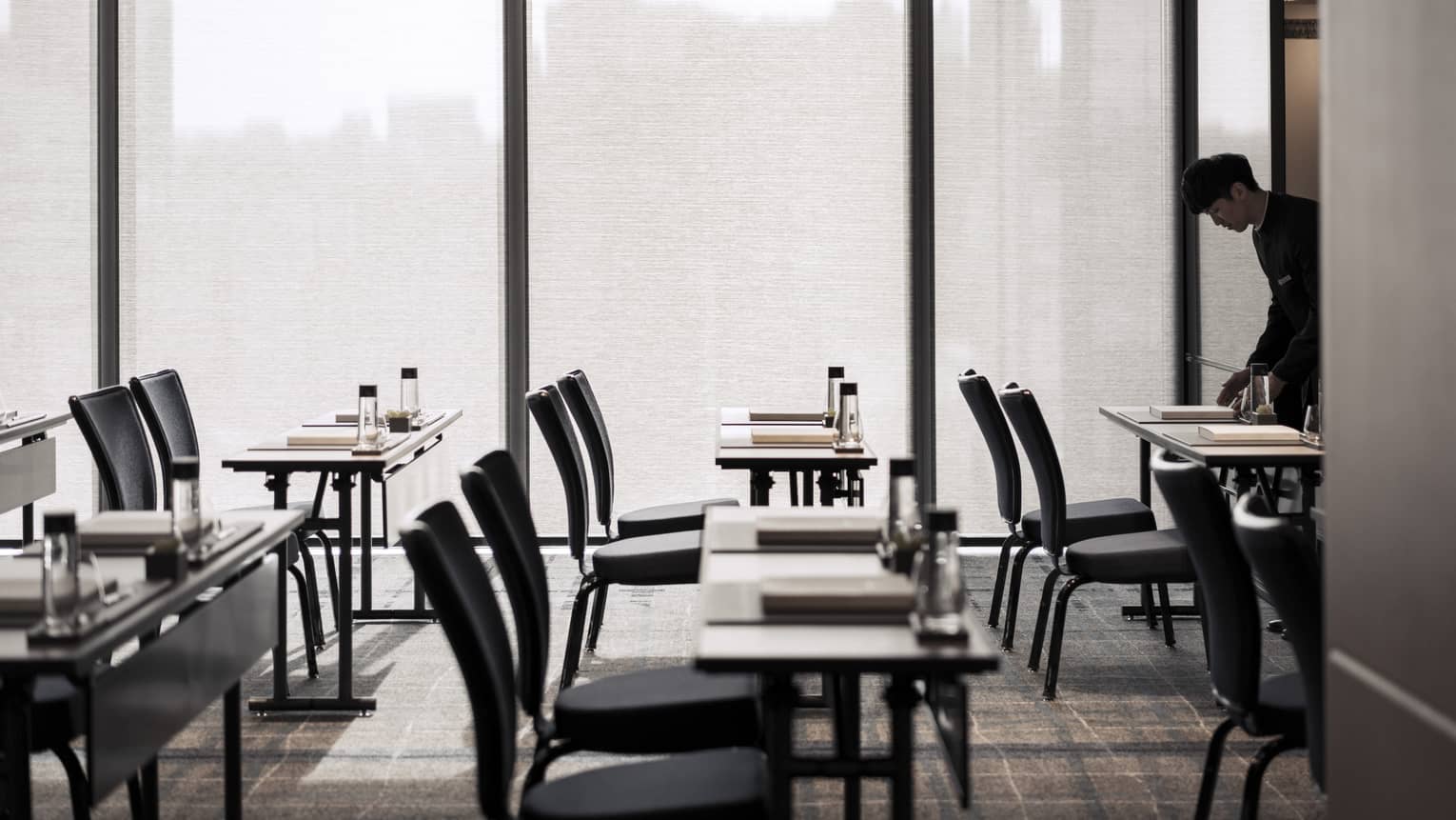 Man setting up rows of desks with water and notebooks in room with large windows